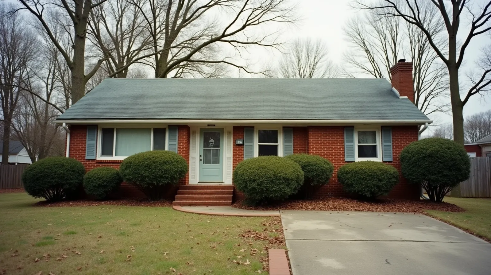 A modest 1970s brick ranch house with an overgrown lawn, bare oak trees, and overcast sky in a quiet Tennessee suburb